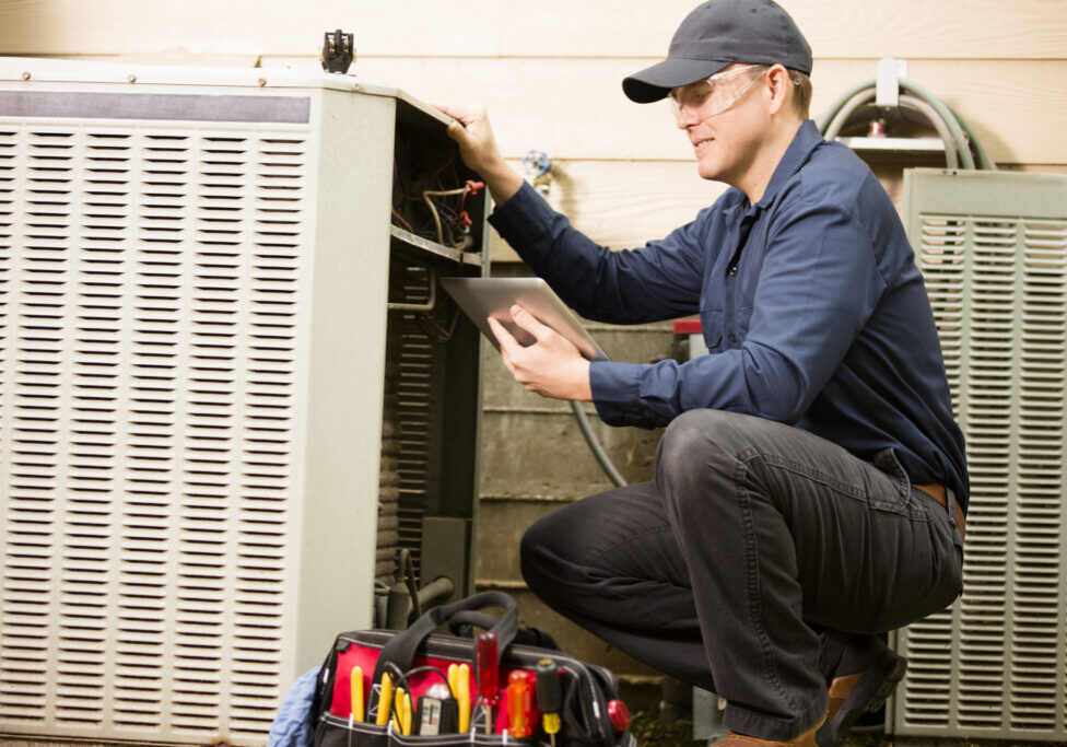 Repairmen works on a home's air conditioner unit outdoors. He is checking the compressor inside the unit using a digital tablet.  He wears a navy blue uniform and his safety glasses.  Tools inside toolbox on ground.